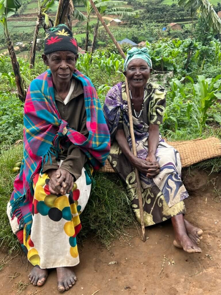 Image 2: An elderly couple sits on a woven bench in a rural setting, surrounded by green plants. The man wears a colorful outfit with a black hat, while the woman holds a wooden stick, dressed in a patterned dress and headscarf. Uganda Amatsiko Tours