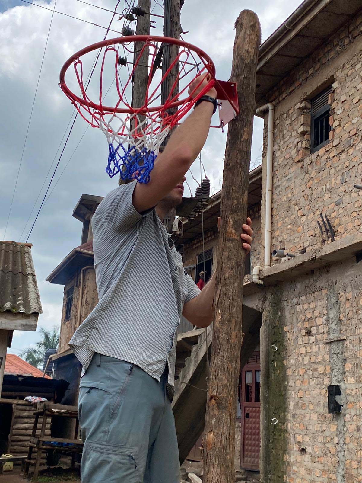 Image 11: A person in a short-sleeved shirt is standing on a ledge, holding a wooden pole with a basketball hoop attached. They're adjusting the hoop outside a rustic brick building under a cloudy sky. Uganda Amatsiko Tours