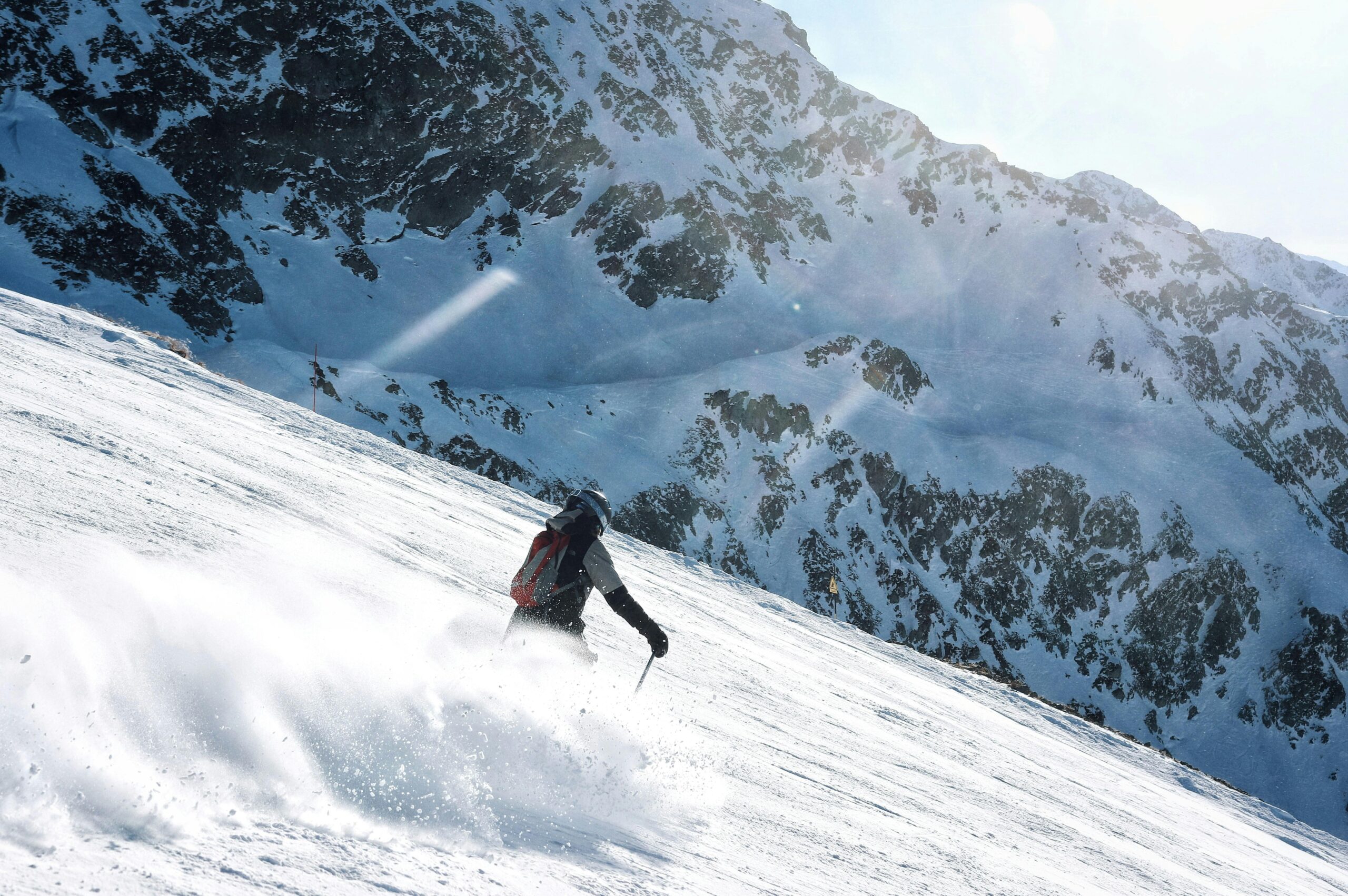 A skier with a red backpack cuts through deep powder snow on a steep alpine slope